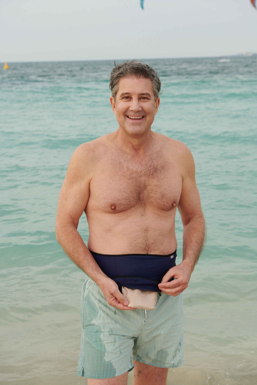 Man at the sea lifting a navy ostomy belt to show his pouch above striped swim shorts
