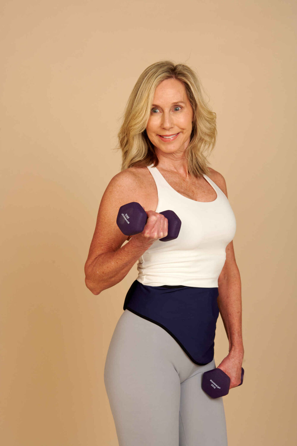 Woman lifting dumbbells in a white tank top and a navy ostomy belt over grey leggings
