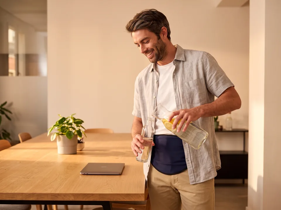 Hydration with an ileostomy: man refilling a water bottle at a co-working table, navy SIIL ostomy belt over his tee